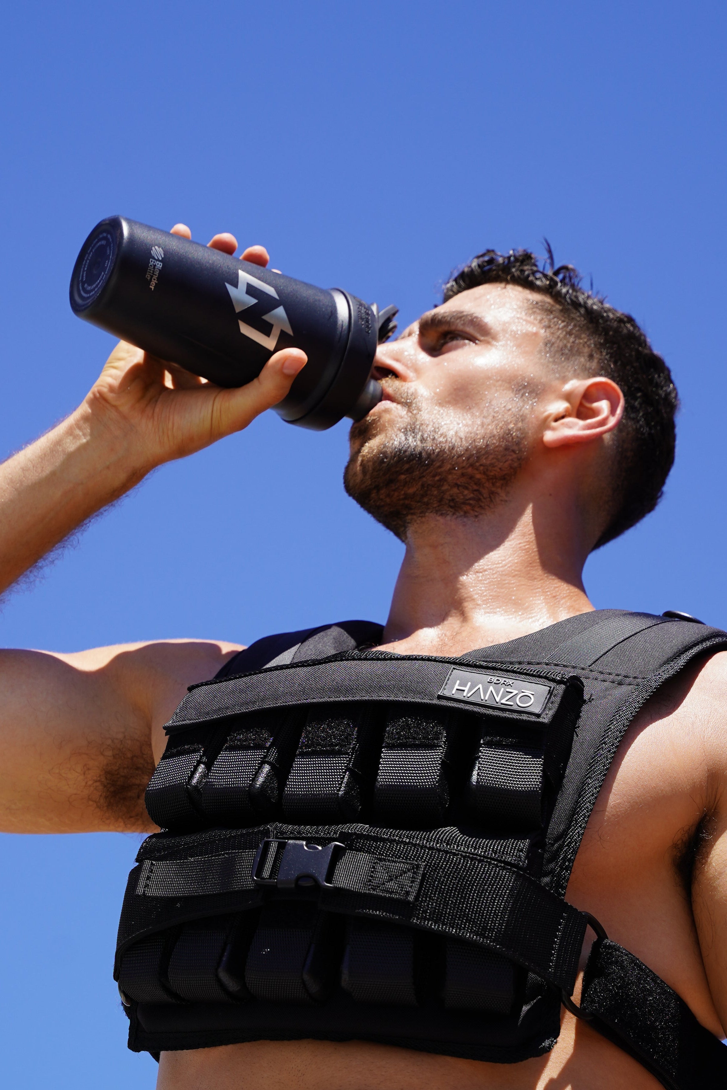 Man drinking from a black water bottle wearing a black weighted vest against a clear blue sky.