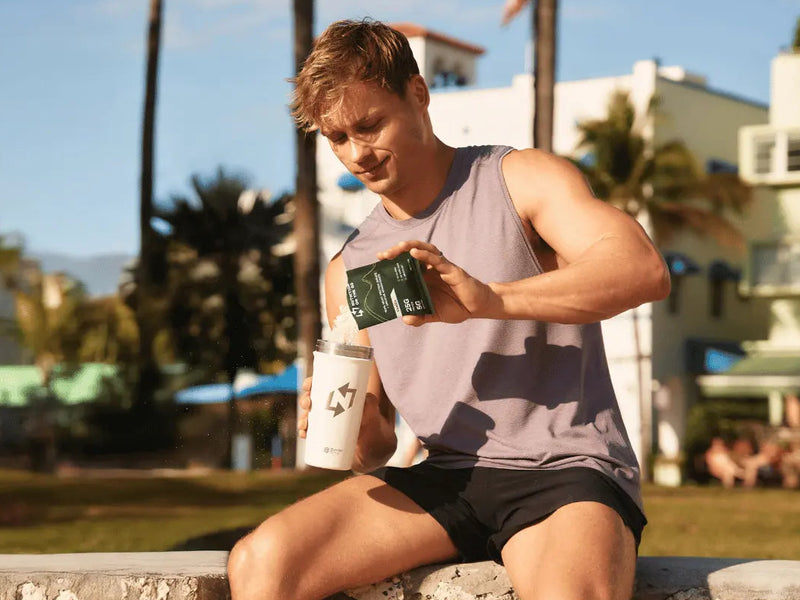 Man sitting on a wall outdoors, holding a phone with palm trees and buildings in the background